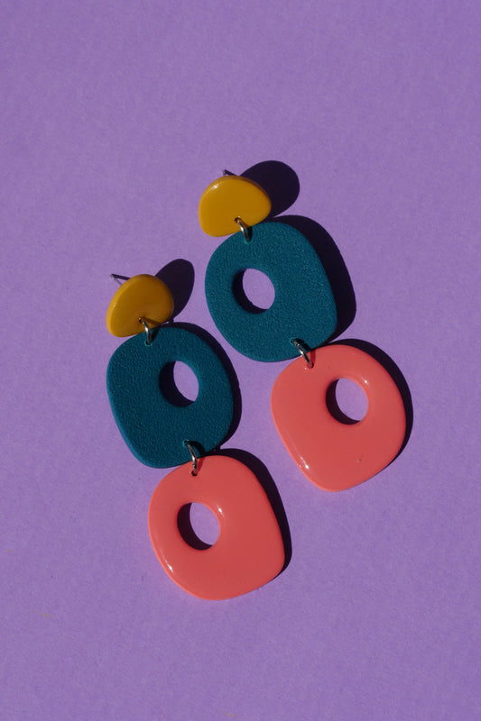 Paire de boucles d’oreilles multicolores en argile polymère posées à plat sur fond violet, formes géométriques aux couleurs vives