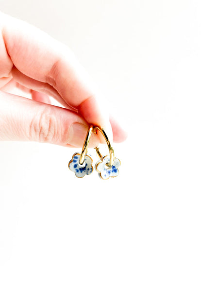 Gros plan des boucles d’oreilles Poppy bleu en forme de fleur, céramique avec détails dorés, tenues entre les doigts