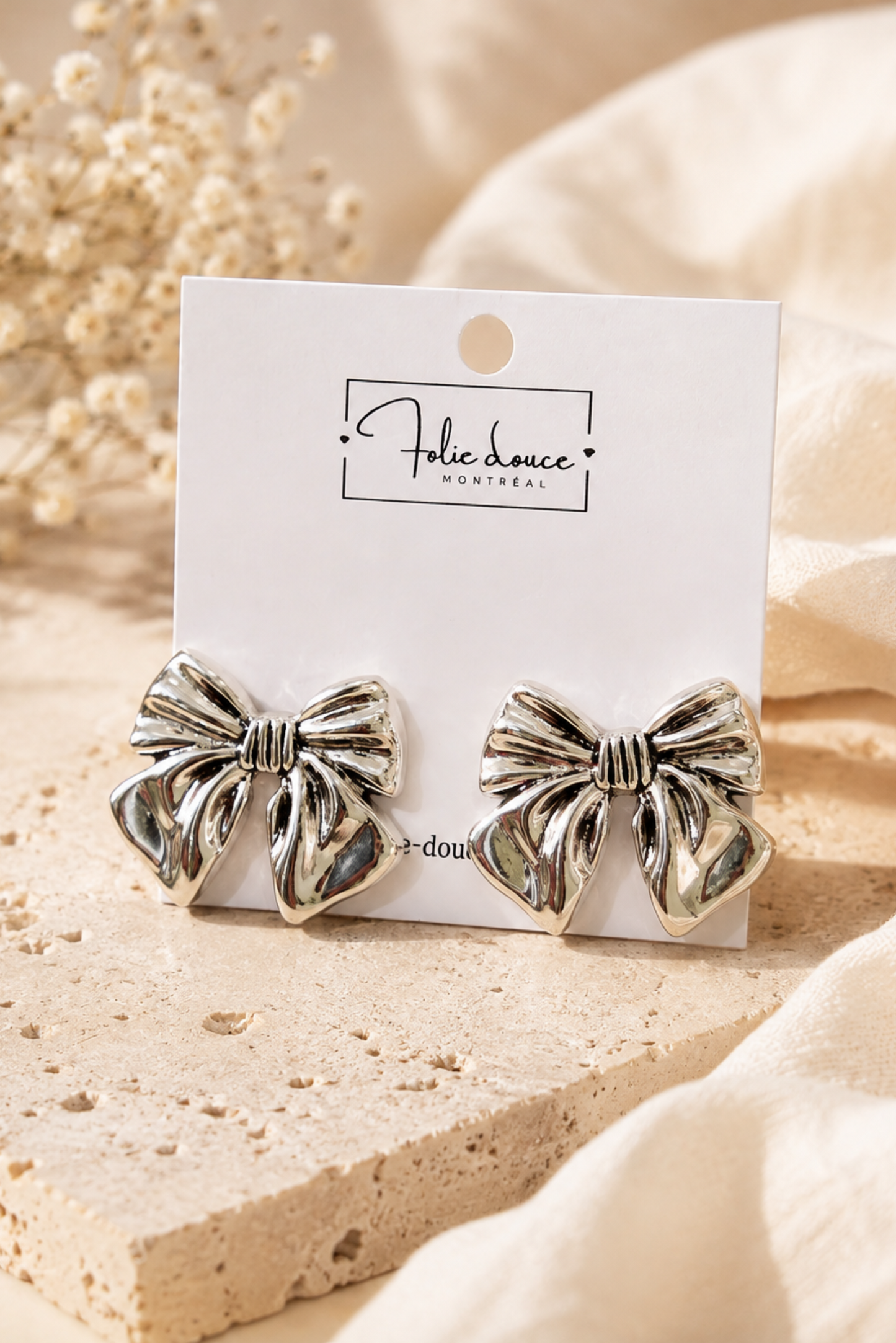 Boucles d’oreilles Nola en forme de nœud argenté, présentées sur une carte Folie Douce avec fleurs séchées.