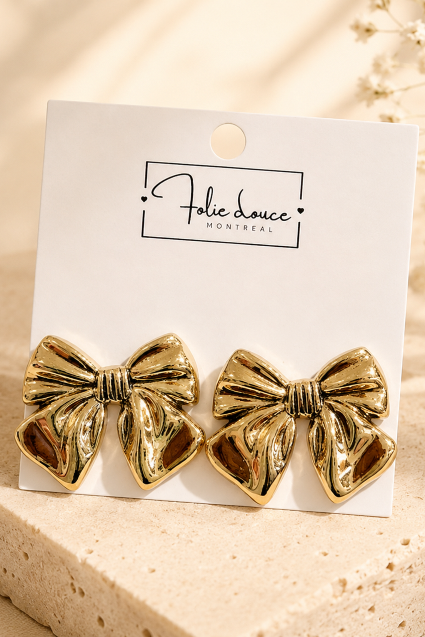 Boucles d’oreilles Nola en forme de nœud doré, présentées sur une carte Folie Douce avec décor doux et lumineux.