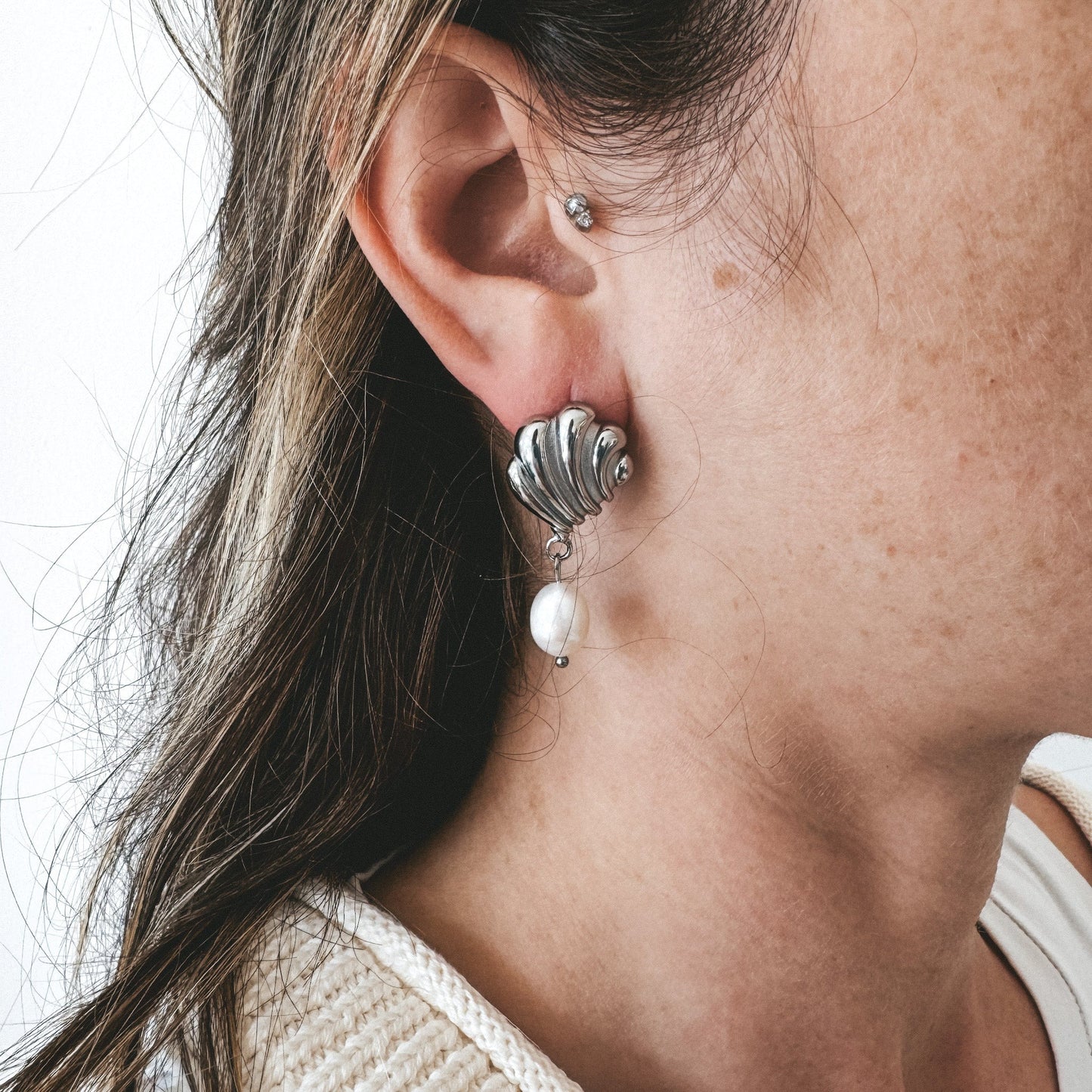 Boucles d'oreilles en acier inoxydable en forme de coquillage avec une perle d'eau douce pendante. Le modèle est de profil et porte une boucle d'oreille cahura et un chandail beige.