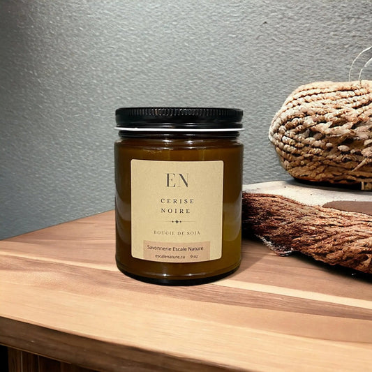 Bougie dans un pot en verre ambre avec un couvercle noir et une étiquette marron.En arrière-plan, on peut voir une table en bois et des décorations en rotin. 