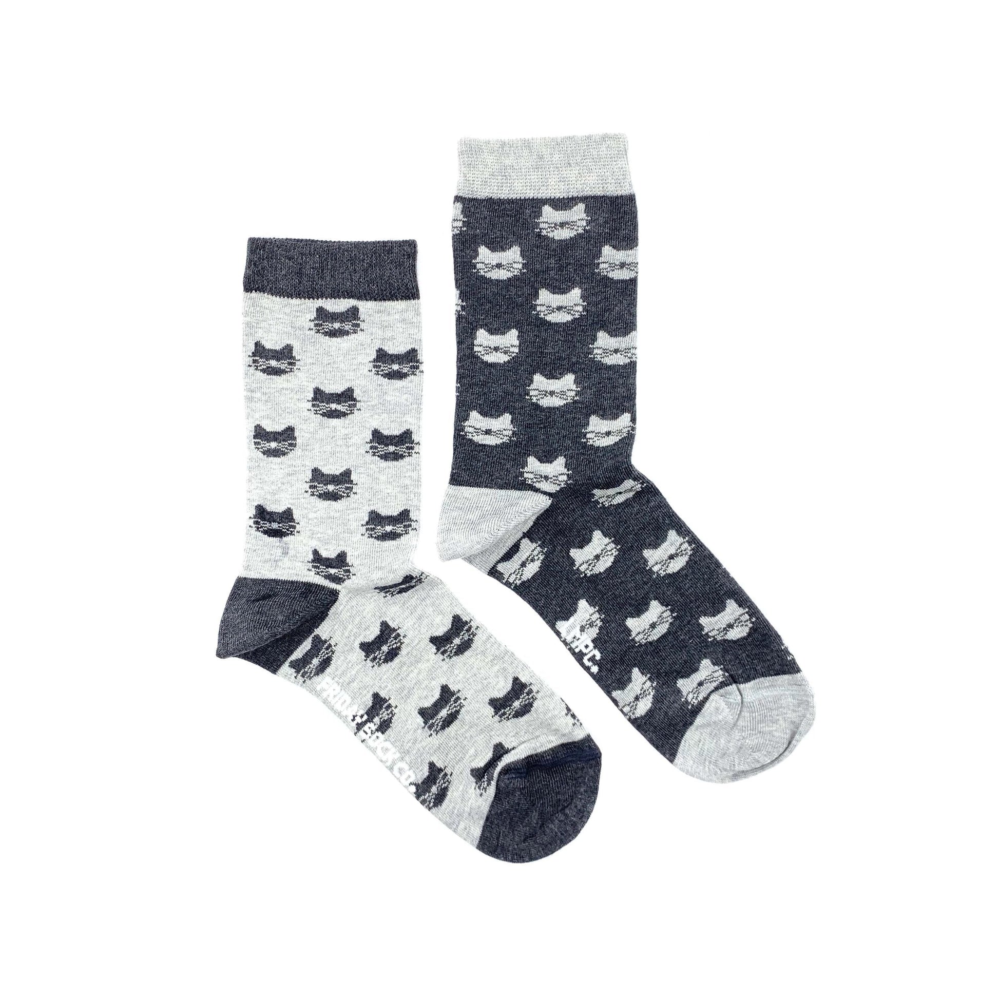 Paire de chaussettes dépareillées sur fond blanc. Une chaussette a les extrémités et le talon gris foncé et des têtes de chat de la même couleur sur le reste de la chaussette. L’autre chaussette a les extrémités et le talon gris clair et des têtes de chat de la même couleur sur le reste de la chaussette. 