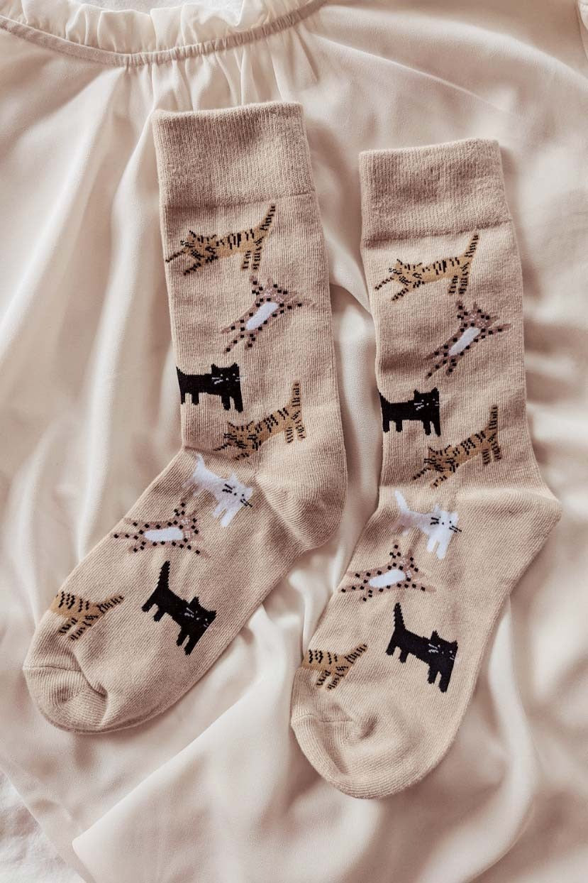 Paire de chaussettes beige avec des chats dans différentes positions. Les chat sont noirs, blancs, tigrés et tachetés. Les chaussettes sont déposés sur un tissu beige. 