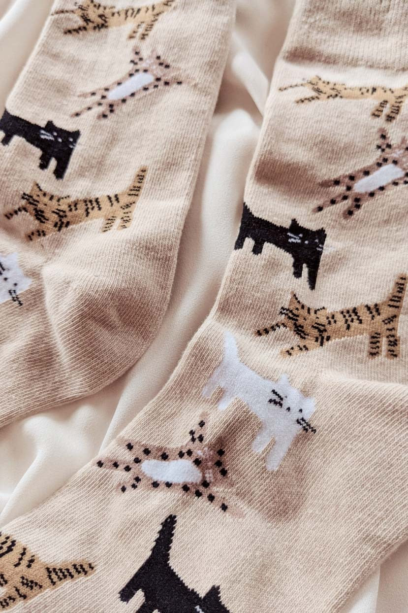 Paire de chaussettes beige avec des chats dans différentes positions. Les chat sont noirs, blancs, tigrés et tachetés. Les chaussettes sont déposés sur un tissu beige. Cette photo est un plan rapproché des chaussettes. 