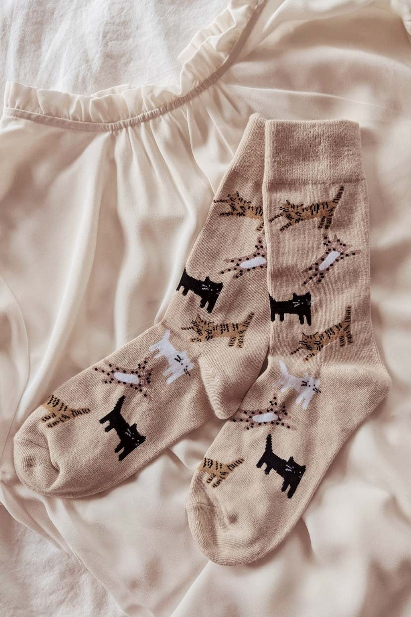 Paire de chaussettes beige avec des chats dans différentes positions. Les chat sont noirs, blancs, tigrés et tachetés. Les chaussettes sont déposés l'une sur l'autre sur un tissu beige. 