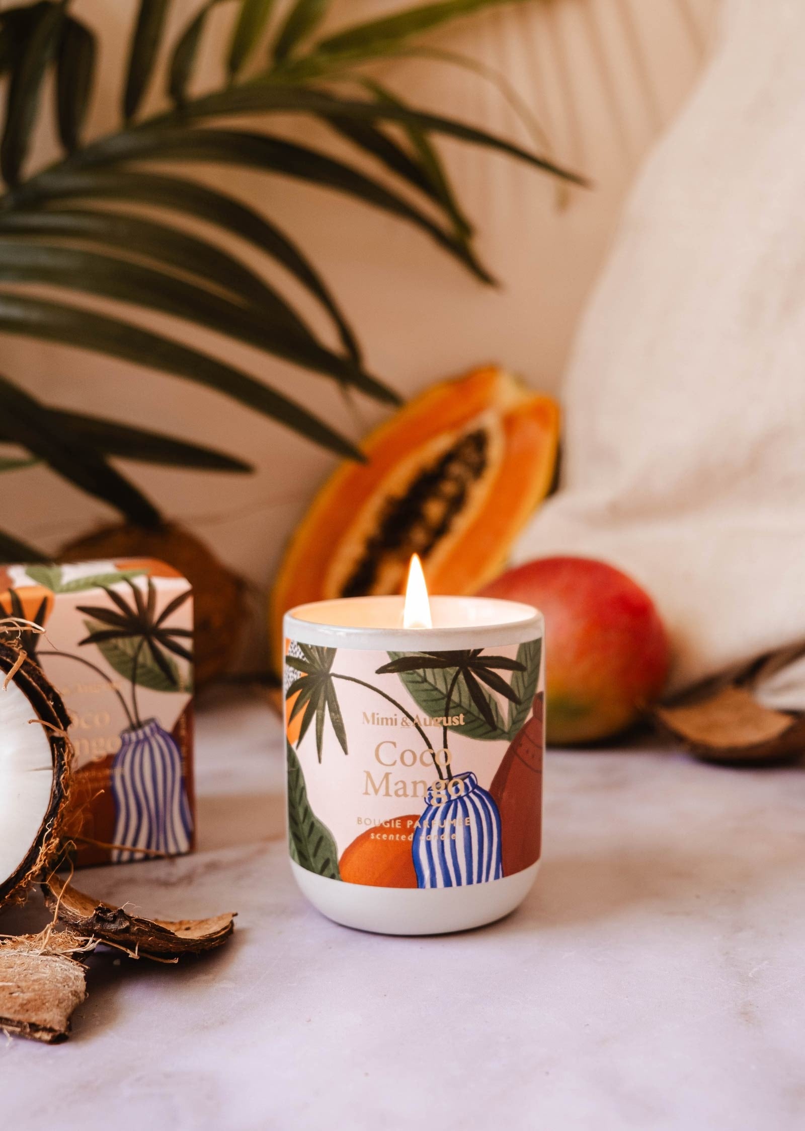 La bougie est coulé dans une tasse en céramique grise sans anse. Le fond de la tasse est arrondi. Sur la bougie, il y a une grosse étiquette sur laquelle on peut lire Mimi & August, Coco Mango, bougie parfumée, scented candle. Sur le fond de l'étiquette, on peut voir un vase rayé blanc et bleu, un pot en terre cuite et des branches de palmier. En arrière-plan, on voit une feuille de palmier et des fruits tropicaux posé sur un tissu beige.