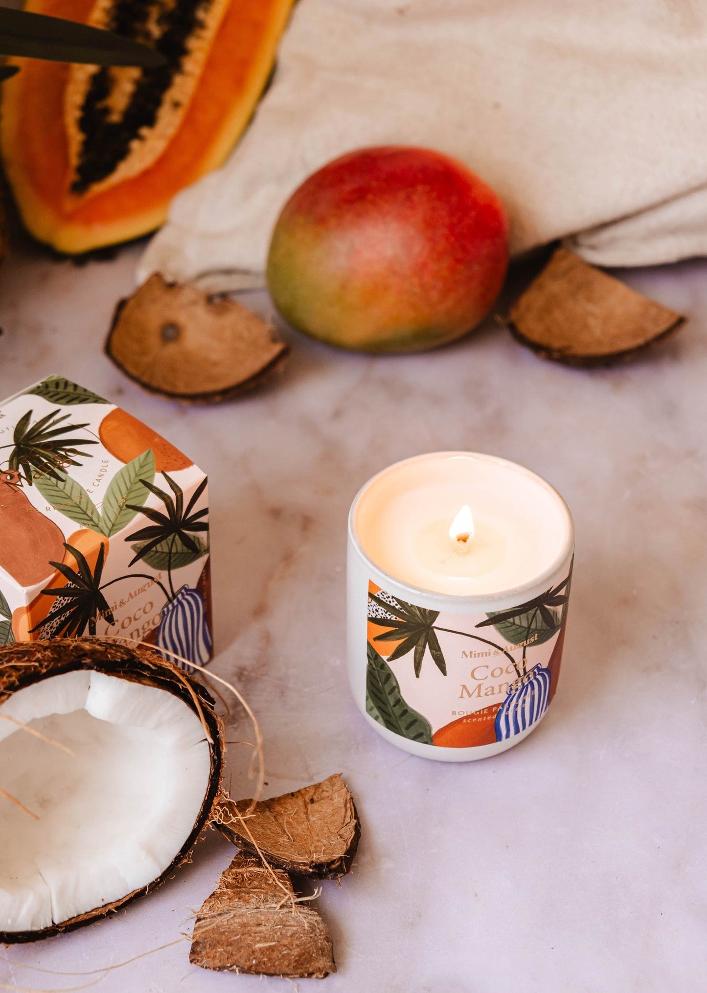 Ici la bougie est posée sur une table en marbre blanc à côté d'une noix de coco, une mangue et une papaye. 