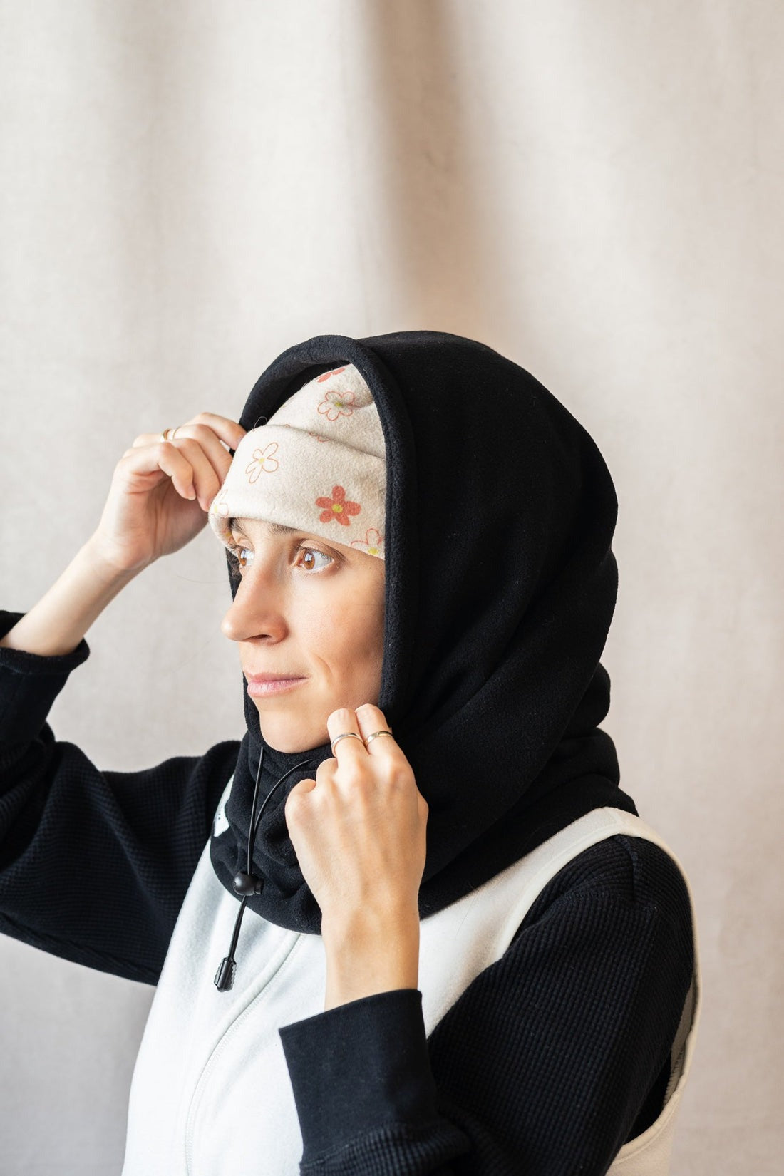 Col capuche avec élastique pour ajuster l'ouverture fait en polaire. Le modèle porte un bonnet beige à fleur en dessous de la capuche et un pull noir et blanc. L'arrière-plan est un fond beige. 