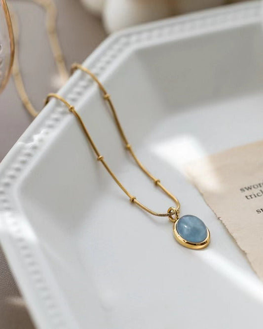 Collier Camelia en acier inoxydable plaqué or 18k avec pendentif ovale et pierre bleutée posé sur assiette blanche