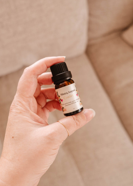 Le flacon d'huile parfumée est une petite bouteille en verre brun sur laquelle est collé une large étiquette beige. Dessus, on peut lire Mimi & August, Confetti, huile parfumée pour diffuseur, scented diffuser oil. Ici, on peut voir quelqu'un tenir le flacon entre son pouce et son index. 