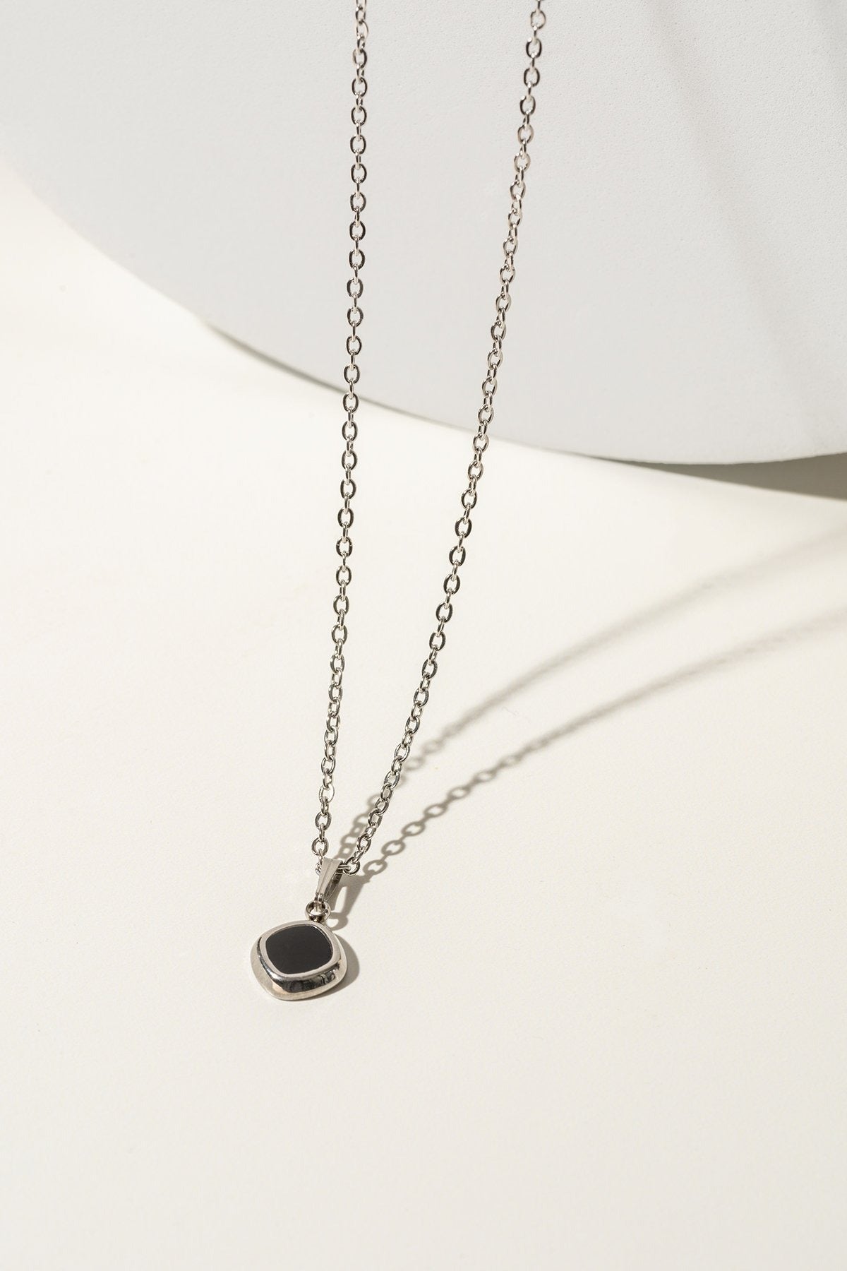 Longue chaine fine en argent avec un pendentif en forme de losange noir. L'arrière-plan est un fond blanc. 