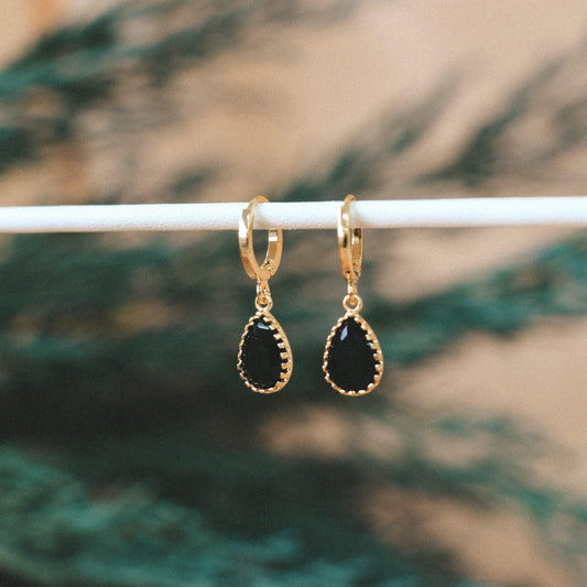 Pendentif en forme de goutte en pierre de verre de couleur noir. L'attache est un anneau doré. Les boucles d'oreilles sont posé sur une tige blanche, l'arrière-plan est un fond vert et beige.