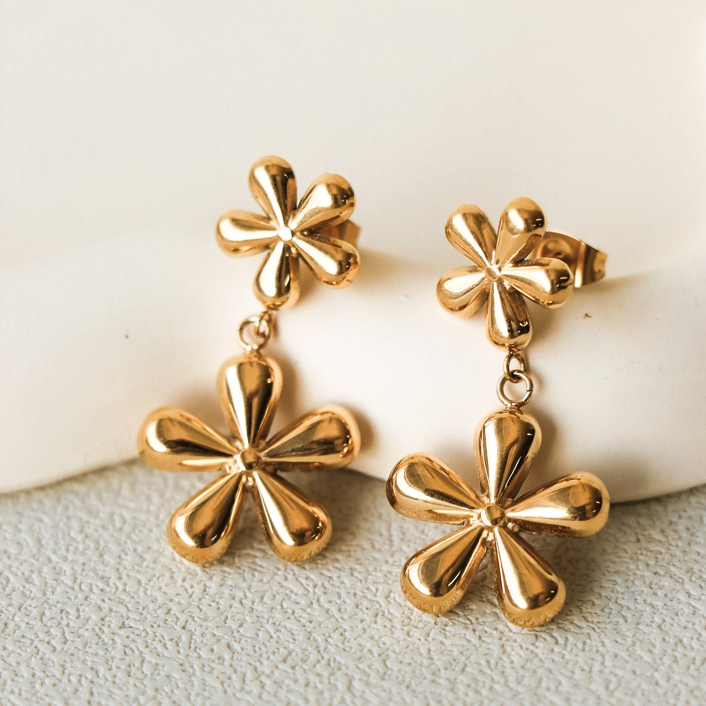 Boucles d'oreilles en forme de fleurs, ce sont deux fleurs de tailles différentes suspendue l'une à l'autre. La paire est faite en acier inoxydable plaqué or. Les boucles d'oreilles sont déposées sur une assiette en céramique beige sur une table grise. Plan rapproché sur les boucles d'oreilles. 