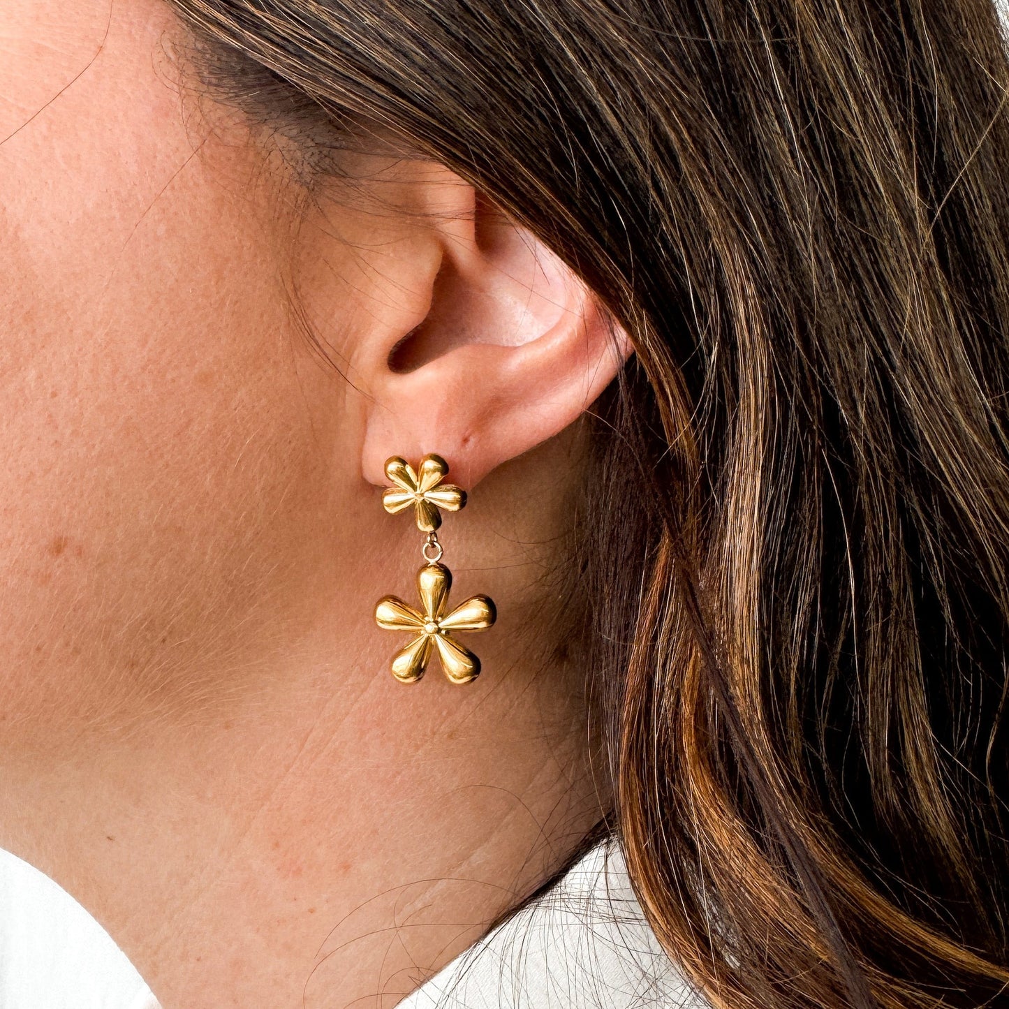 Boucles d'oreilles en forme de fleurs, ce sont deux fleurs de tailles différentes suspendue l'une à l'autre. La paire est faite en acier inoxydable plaqué or. Les boucles d'oreilles sont portées par un modèle qui se tient de profil.