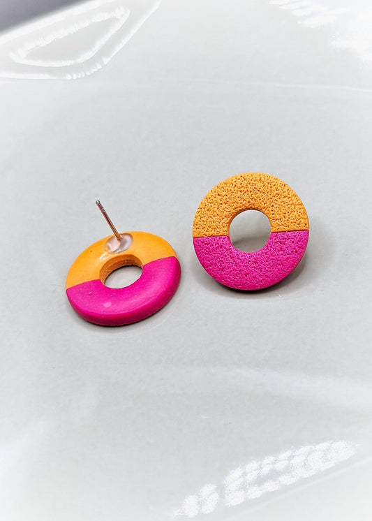 Petites boucles d'oreilles rondes avec un trou au milieu. Elles sont texturées avec un papier ponce qui leur donne un aspect de pierre. Elles sont divisées en deux couleurs, la partie en haut est orange et celle d'en bas magenta. Une des boucles d'oreilles est à l'envers, on peut voir que le derrière est vernis. Les boucles d'oreilles sont présentées sur une assiette blanche. 
