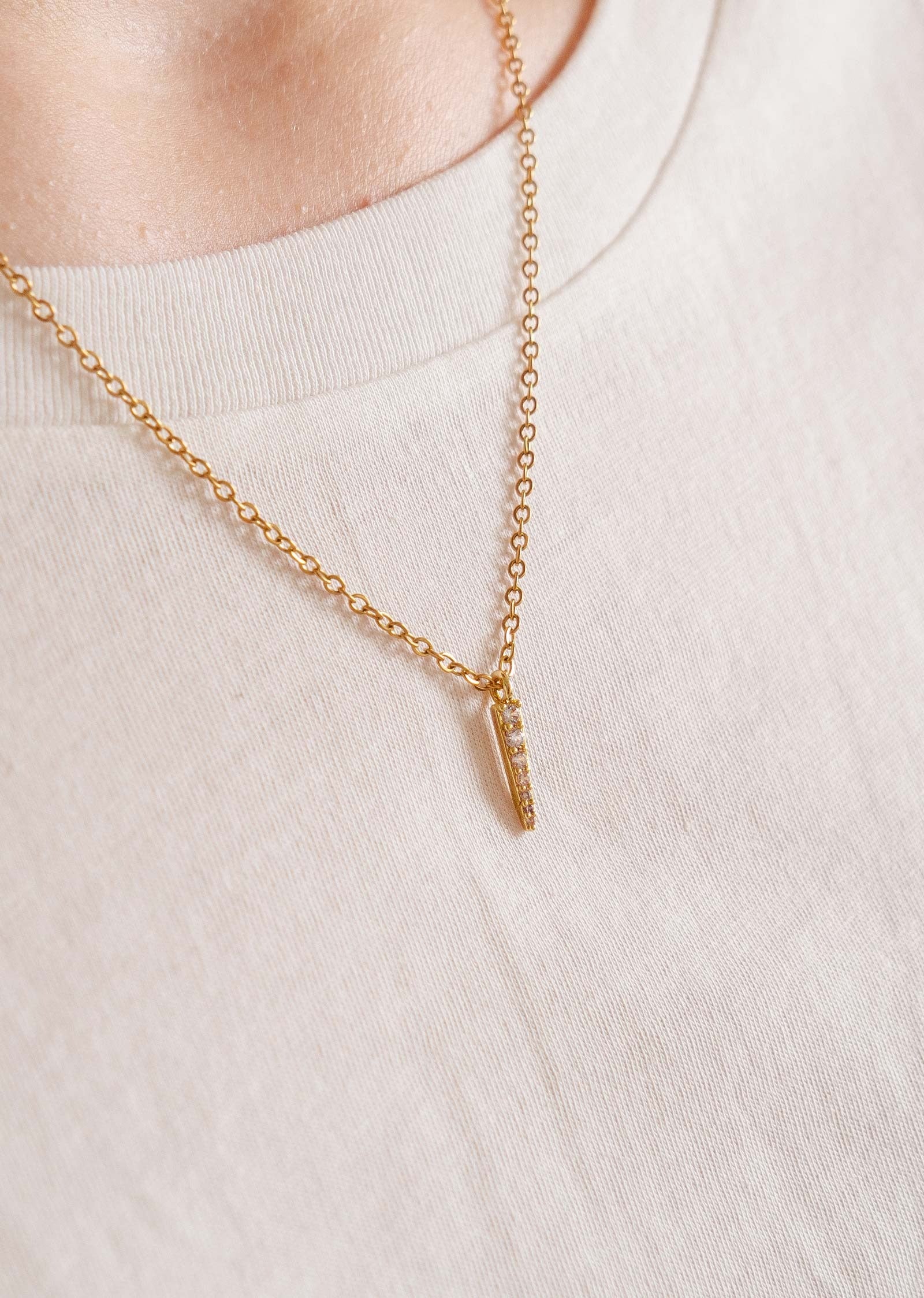 Fine chaîne dorée avec un pendentif en forme de barre orné de zircons scintillants. On peut voir que quelqu'un porte le collier autour de son coup. La personne se tient sur le côté et porte un t-shirt beige. 