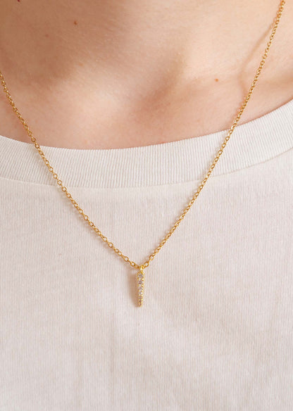 Fine chaîne dorée avec un pendentif en forme de barre orné de zircons scintillants. On peut voir que quelqu'un porte le collier autour de son coup. La personne est de face et porte un t-shirt beige. 