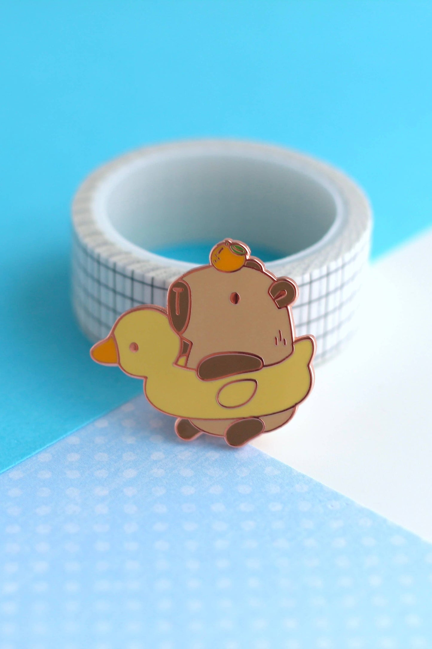 Épinglette Capybara et Canard en émail dur plaqué cuivre, posée sur fond pastel bleu et blanc.