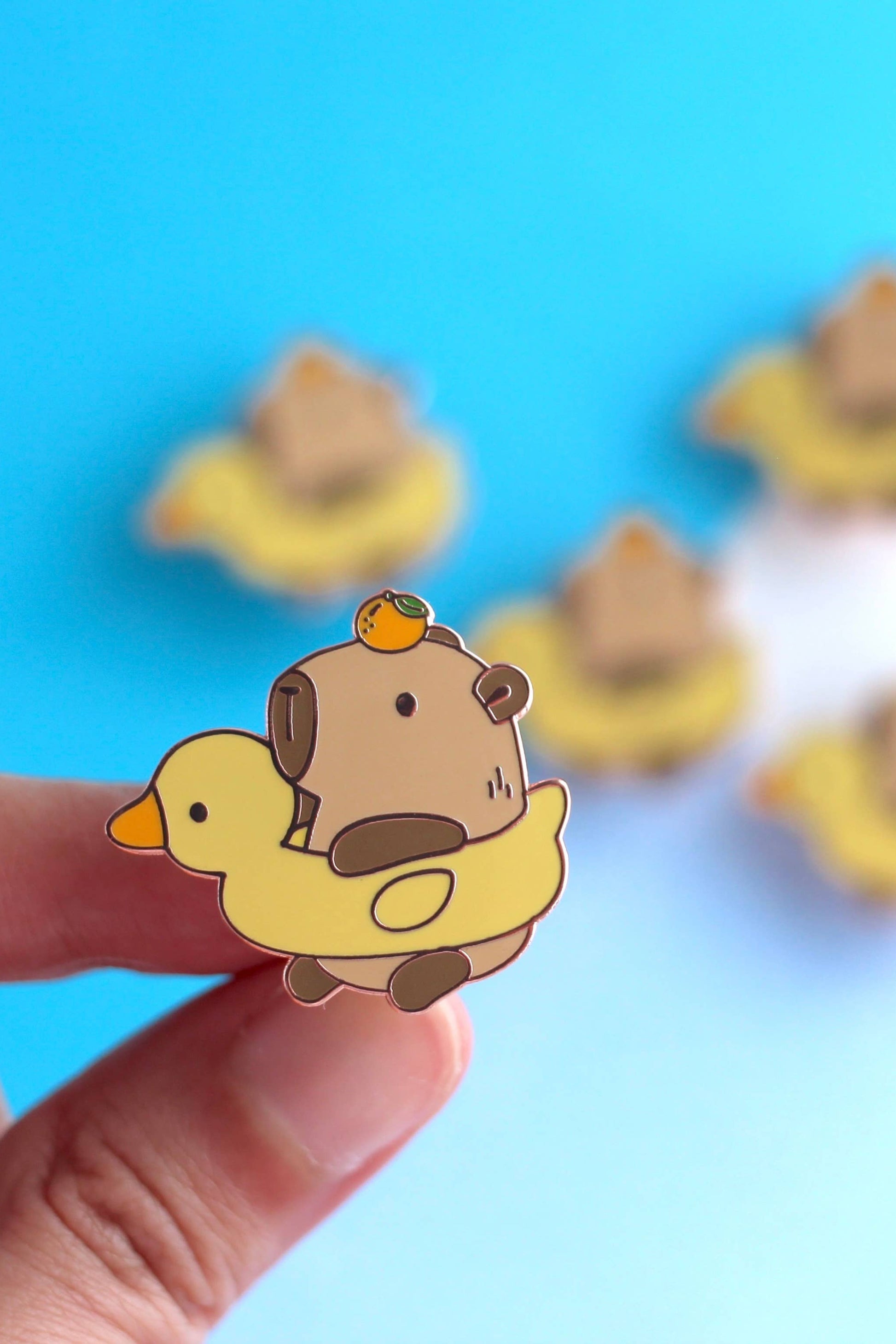 Épinglette Capybara et Canard en métal cuivré, tenue entre les doigts sur fond bleu pastel.