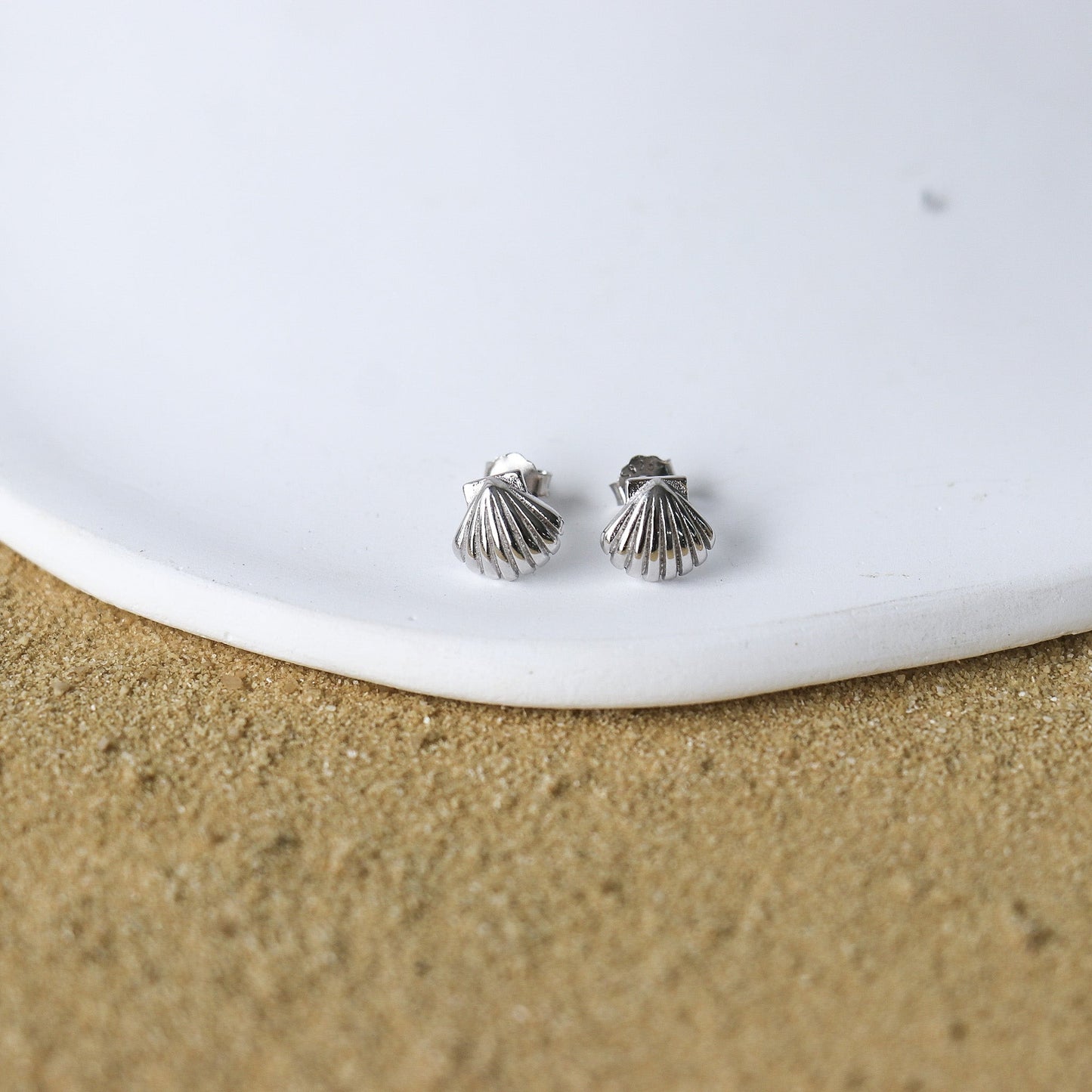 Clous d'oreilles délicats en forme de petit coquillage fait en argent .925. Les boucles d'oreilles sont déposé sur une assiette blanche elle-même posé sur du sable.