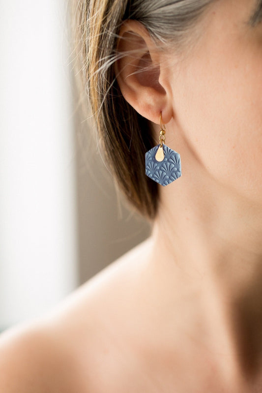 Boucles d'oreilles en argile polymère bleu en forme d'hexagone avec une petite plaque en laiton en forme de goute. Le modèle se tient de profil, c'est un plan rapproché. 