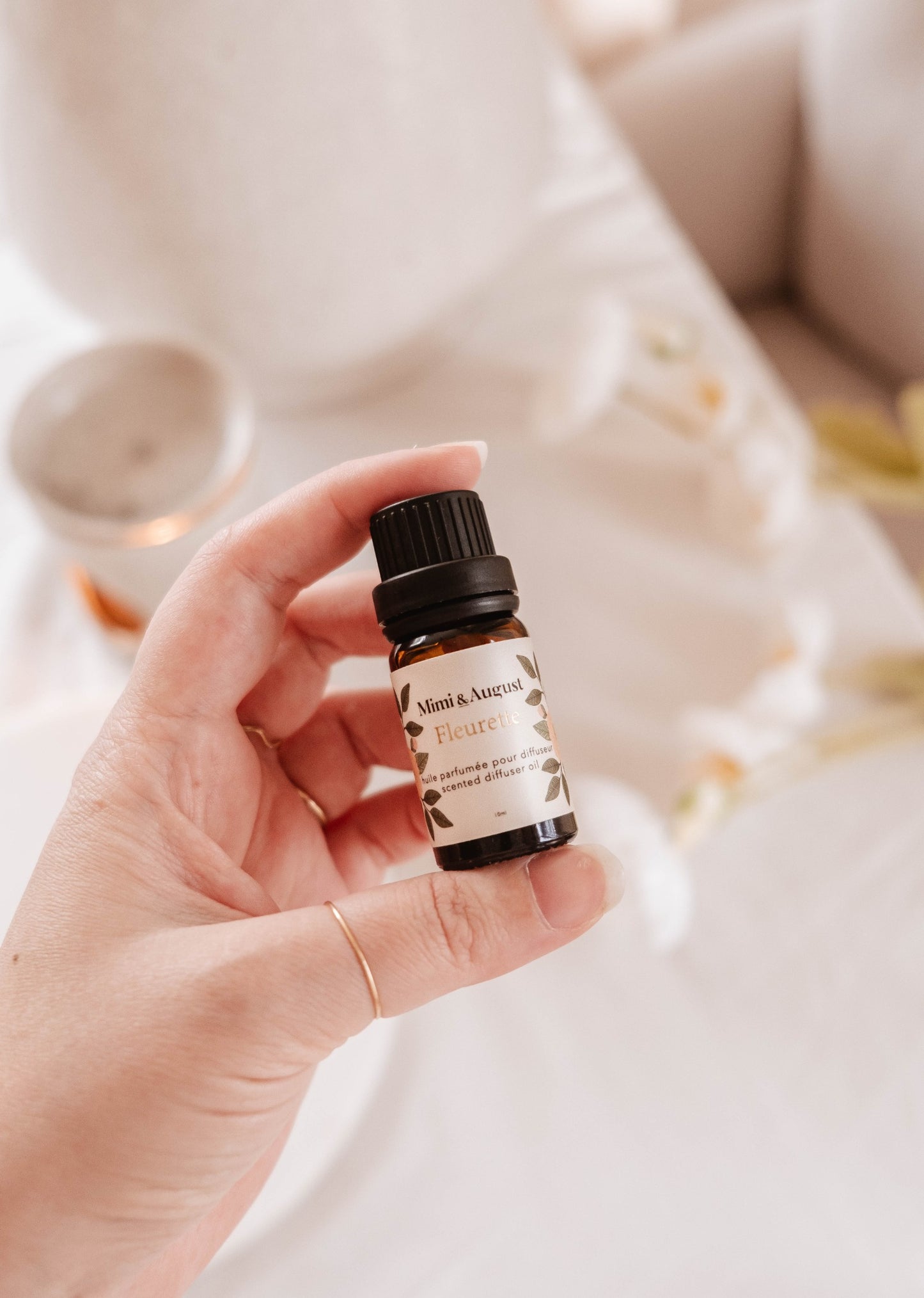 Le flacon d'huile parfumée est une petite bouteille en verre brun sur laquelle est collé une large étiquette beige. Dessus, on peut lire Mimi & August, Fleurette, huile parfumée pour diffuseur, scented diffuser oil. Ici, on peut voir quelqu'un tenir le flacon entre son pouce et son index. 