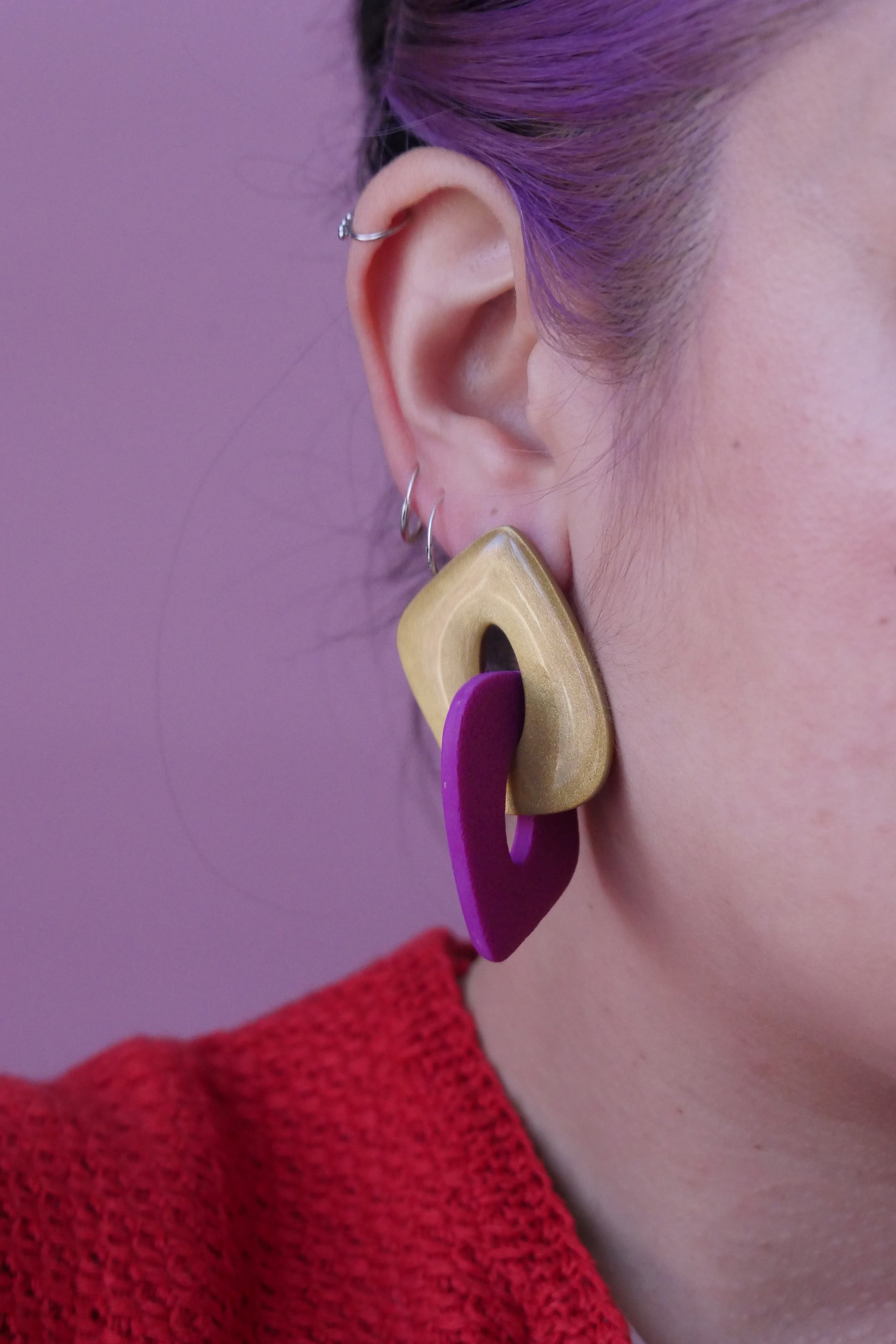 Les boucles d'oreilles sont faites de deux triangles creux et arrondies qui s'entremêlent. Un est à paillettes dorées et l'autre fushia. Plan rapproché sur l'oreille d'un modèle portant une boucle d'oreilles et trois petits anneaux fins argent. 