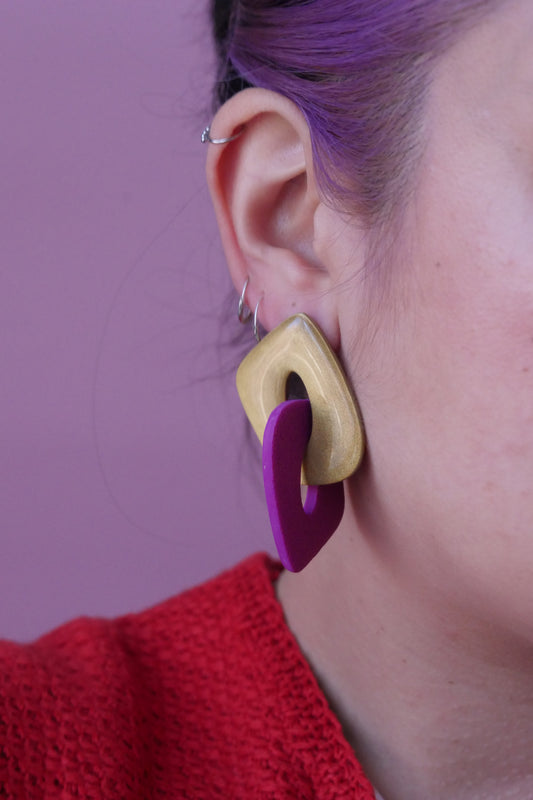 Les boucles d'oreilles sont faites de deux triangles creux et arrondies qui s'entremêlent. Un est à paillettes dorées et l'autre fushia. Plan rapproché sur l'oreille d'un modèle portant une boucle d'oreilles et trois petits anneaux fins argent. 