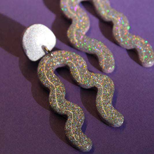 Longues boucles d'oreilles en forme de zig-zag à paillettes argent aux reflets irisé. L'arrière-plan est un fond violet. 