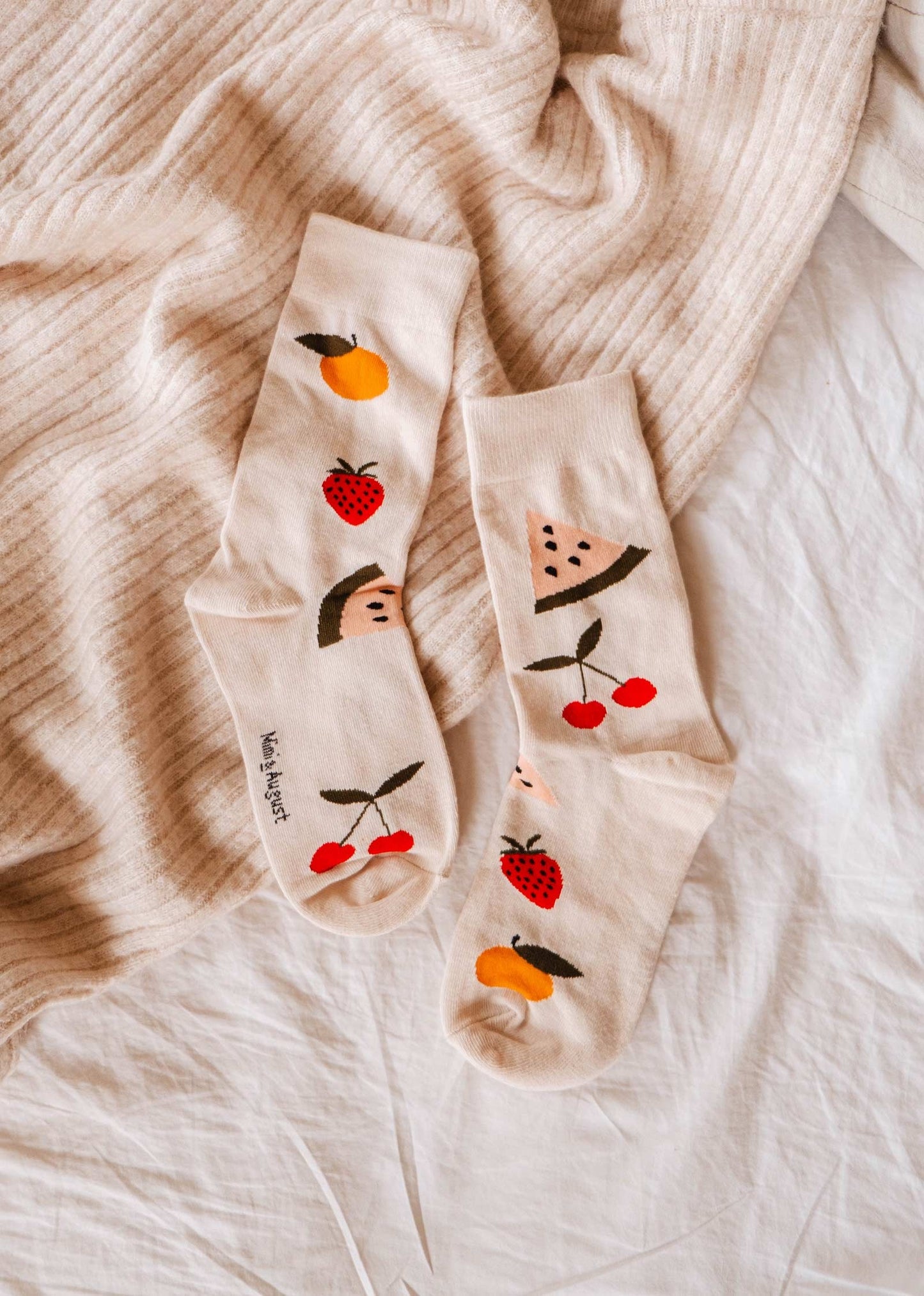 Paire de chaussettes mi-haute beige avec des gros fruits; cerises, fraises, pêches et melon d'eau. Cette photo est un plan éloigné des chaussettes qui sont déposés sur des tissus de textures différentes dans des tons beige. 