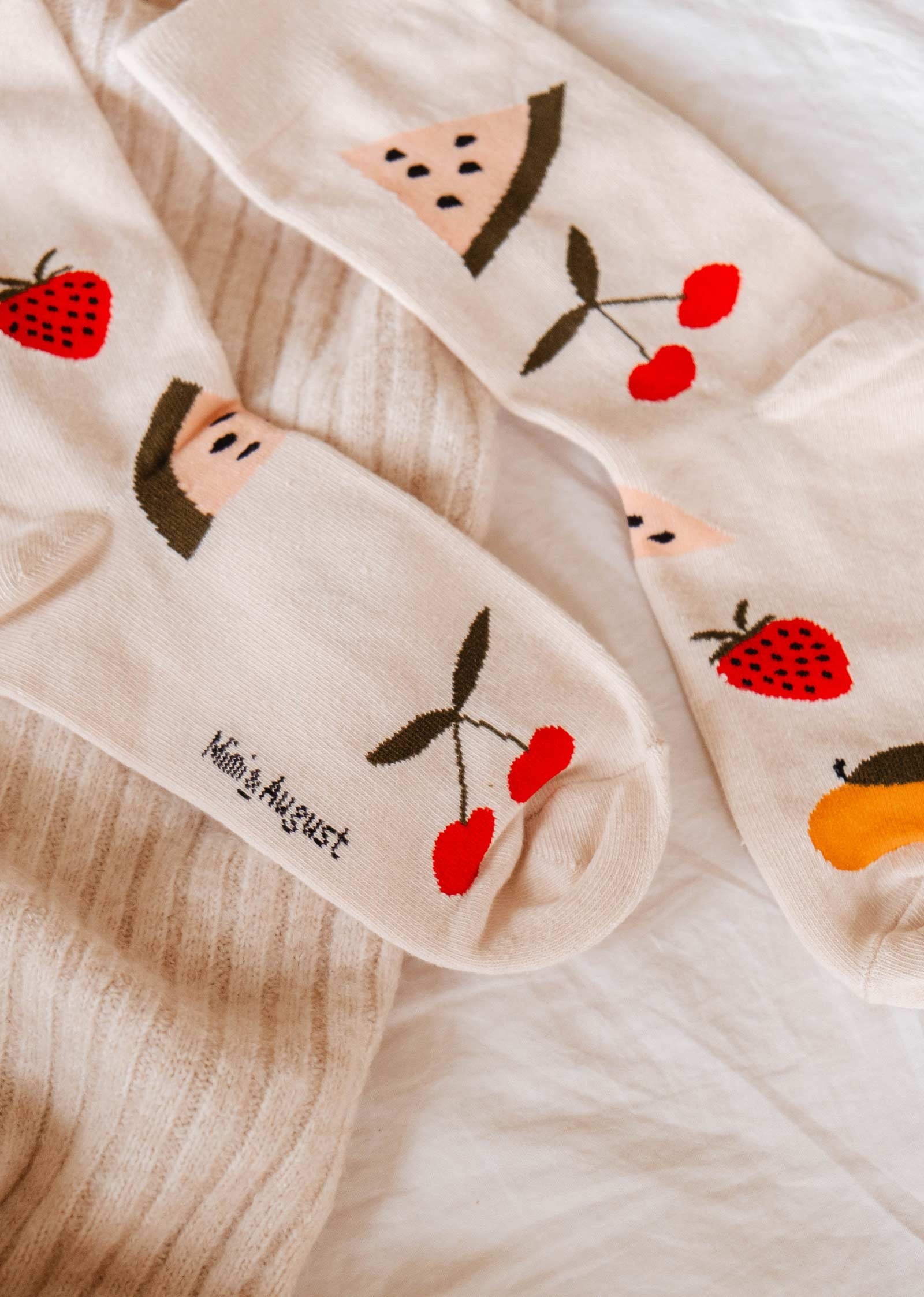 Paire de chaussettes mi-haute beige avec des gros fruits; cerises, fraises, pêches et melon d'eau. Cette photo est un plan rapproché des chaussettes qui sont déposés sur des tissus de textures différentes dans des tons beige. 