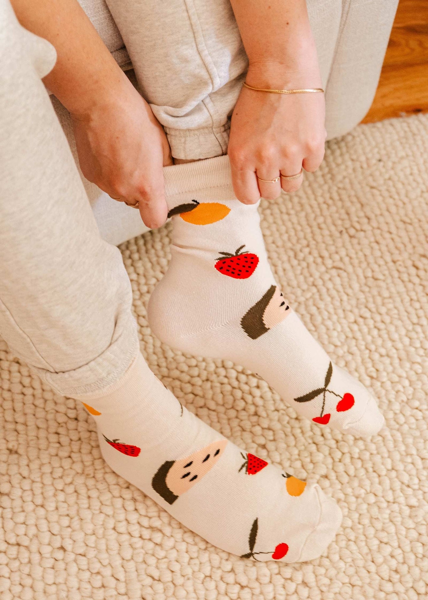 Paire de chaussettes mi-haute beige avec des gros fruits; cerises, fraises, pêches et melon d'eau. Sur cette photo, on peut voir quelqu'un mettre les chaussettes, elle est assise sur un canapé beige et en-dessous de ses pieds il y a un tapis beige. 