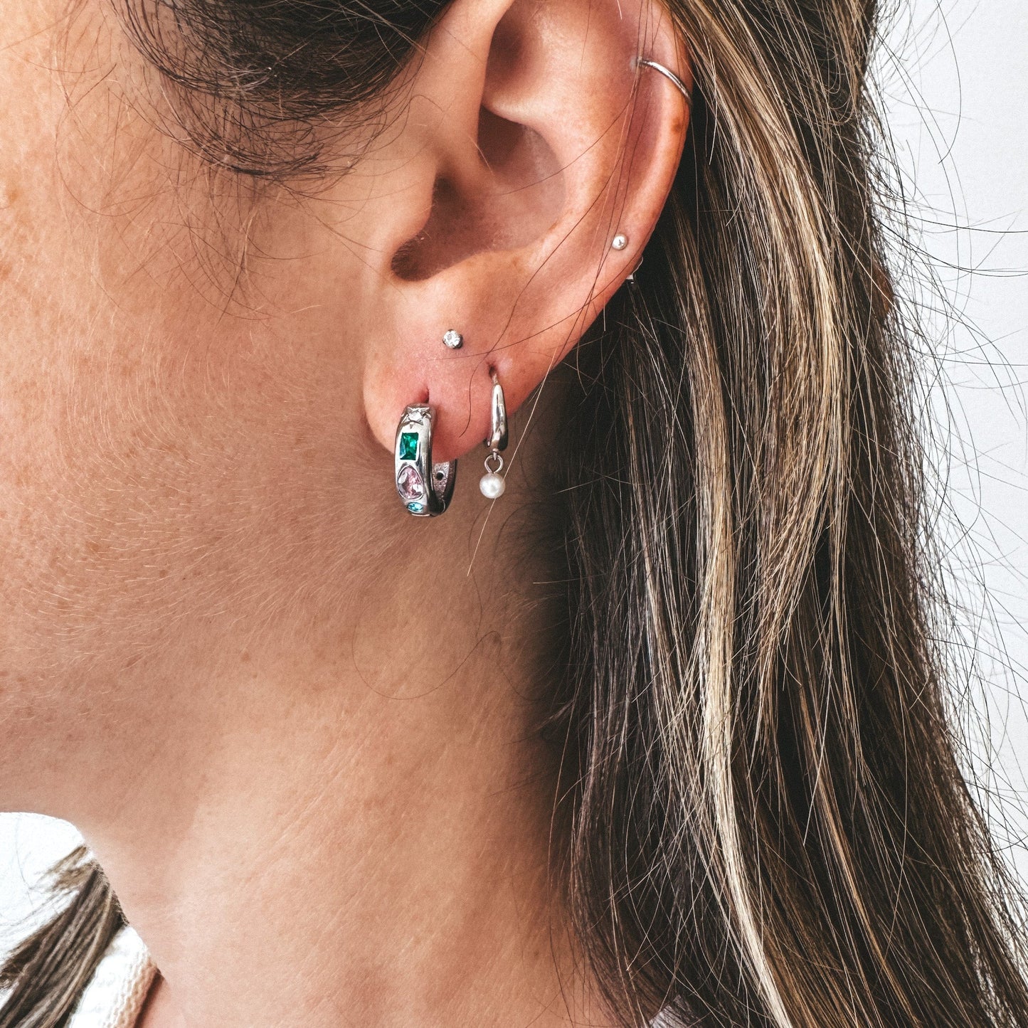 Anneaux argent en acier inoxydable avec petits zircons vert, rose, blanc et bleu et de forme variées. Les boucles d'oreilles sont portées par un modèle qui se tient de profile. La personne porte d'autres boucles d'oreilles argent de la même marque; un anneau argent avec une perle d'eau douce en pendentif et un petit clou d'oreilles avec un zircon blanc. 