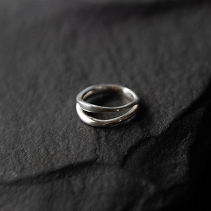 Bague argent minimaliste avec un design entrecroisé. Les deux bandes fines se croisent délicatement, créant un effet de mouvement fluide et élégant. Le fond en pierre noire texturée met en valeur la finition brillante et raffinée du métal argent.