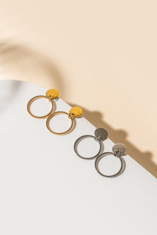 La partie sur l'oreille est un tout petit disque auquel est suspendu un anneau très fin. Sur la photo on peut voir la paire de boucles d'oreilles en version argent et en version or posées sur le bord d'un plateau blanc. L'arrière-plan est beige. 