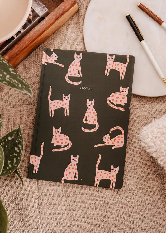 La couverture du carnet de notes est verte foncé avec des petits chats rose tachetés dans plein de positions rigolotes. Le carnet est déposé sur une nappe beige. On voit autour une plante, des livres et des crayons.