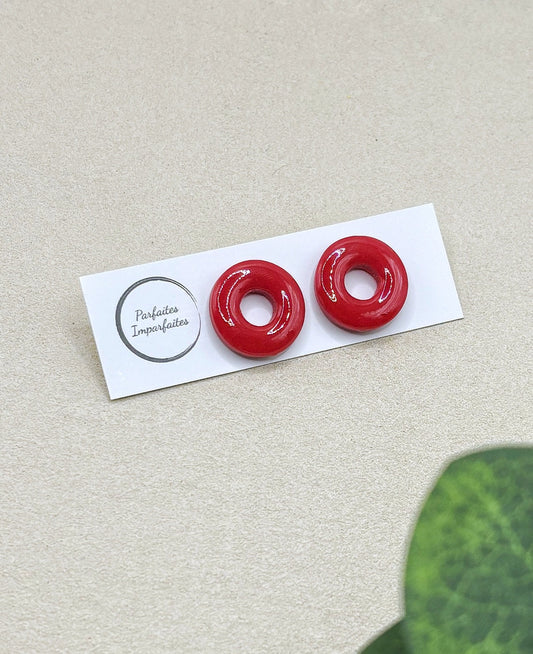 Petites boucles d'oreilles ronde avec un trou au centre. Elles sont vernis ce qui leur donne vraiment l'apparence de bonbons. Elles sont entièrement rouge. Les boucles d'oreilles sont présentées sur un carton blanc avec le logo de la marque Parfaites Imparfaites et l'arrière-plan est un fond beige. 