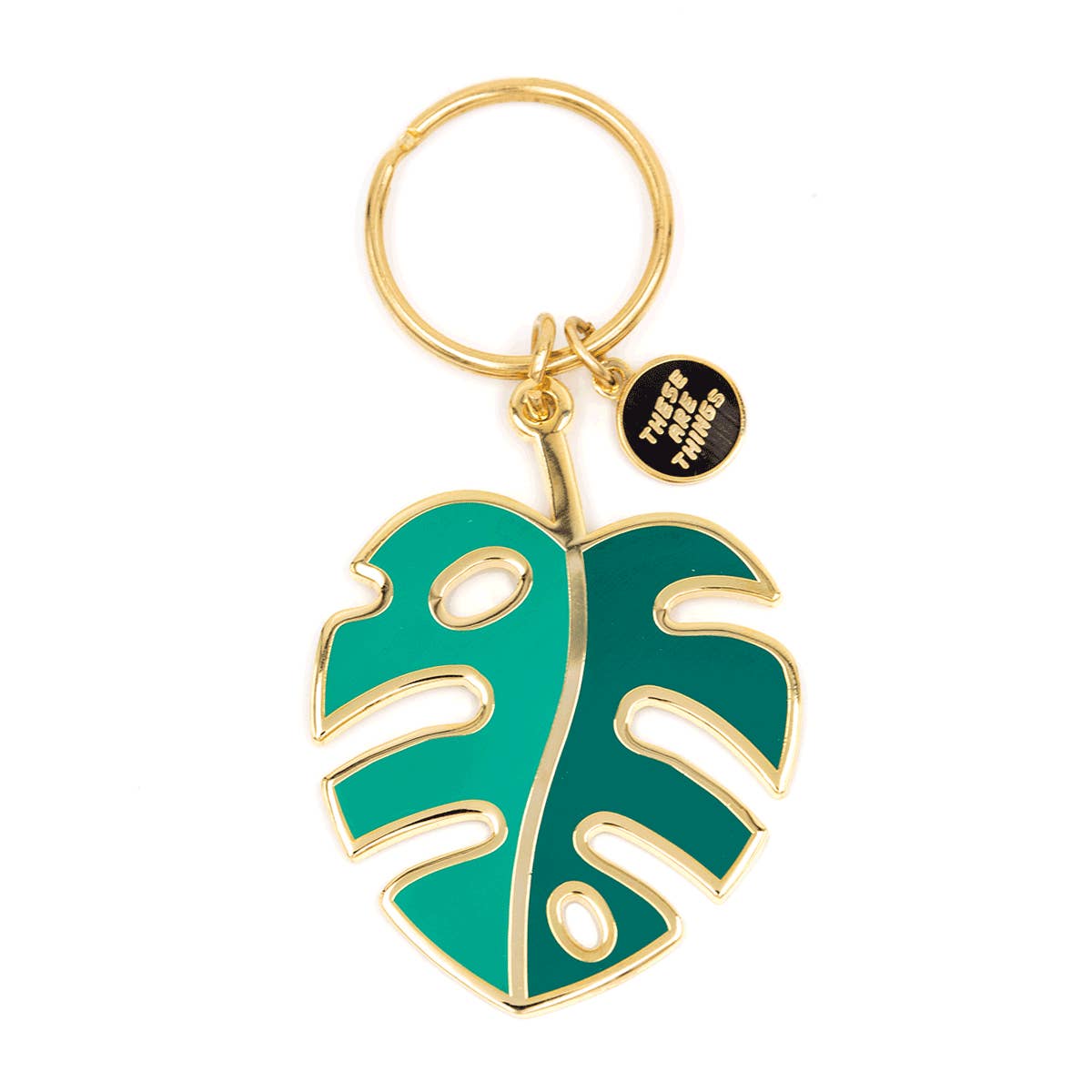 Porte-clef et breloque en forme de feuille de monstera. Le porte-clef est en émail de couleur or et la feuille verte. Il y a une toute petite breloque sur laquelle est inscrit le nom de la marque qui est There are things. L’arrière-plan est un fond blanc. 