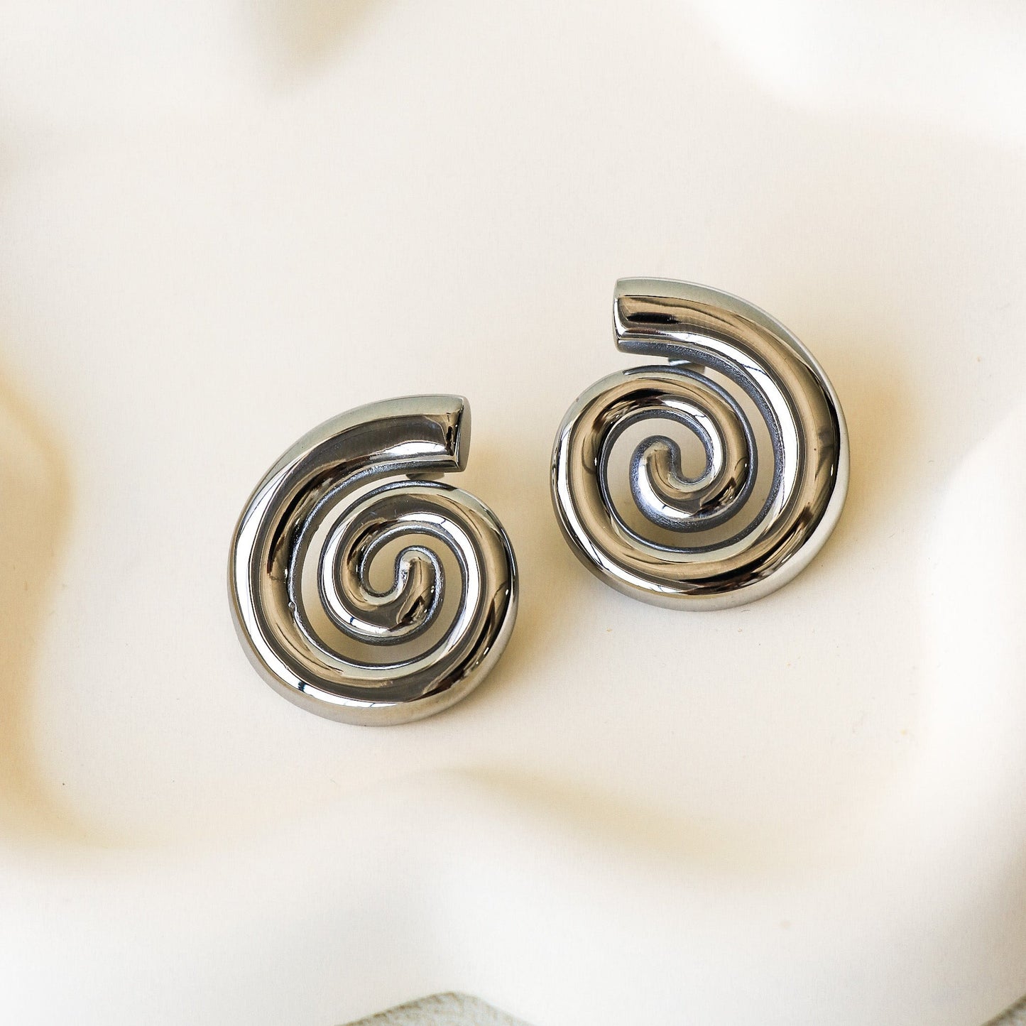 Boucles d'oreilles argent en acier inoxydable en forme de spirale. Les boucles d'oreilles sont posées sur un fond beige.