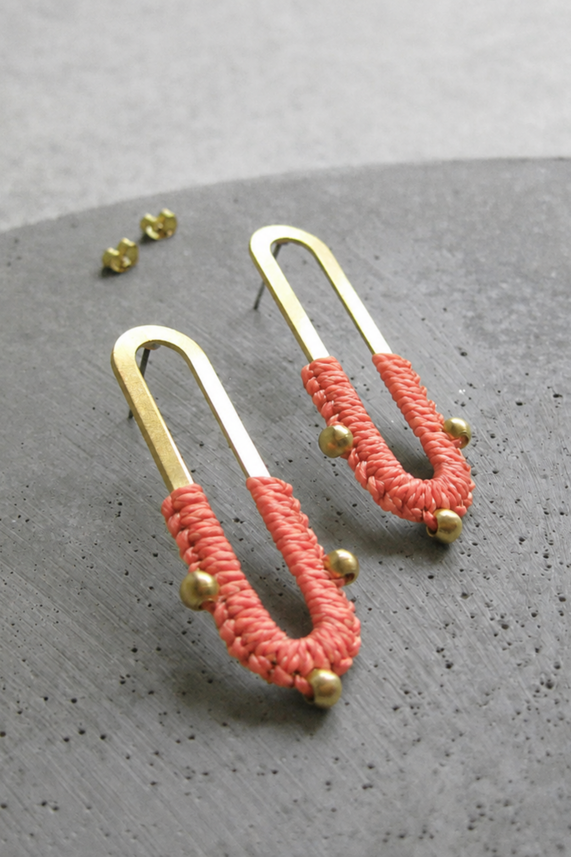 Boucles d’oreilles Nyx Corail posées sur fond gris, design en laiton doré avec fibre textile corail.