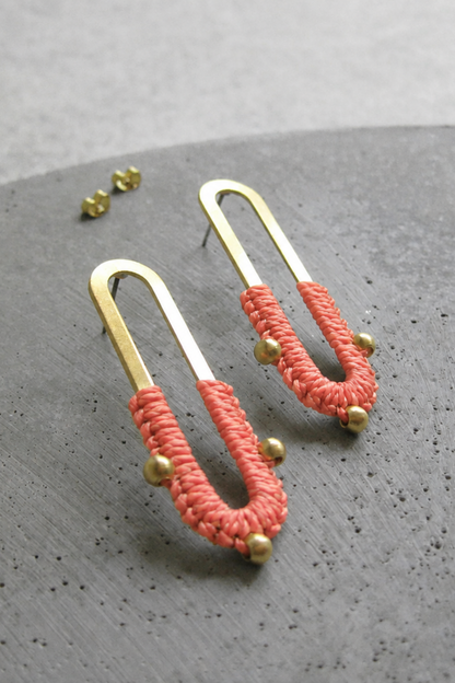 Boucles d’oreilles Nyx Corail posées sur fond gris, design en laiton doré avec fibre textile corail.