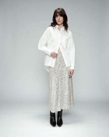 Jupe longue à sequins argentés avec ceinture élastique. Le modèle porte une chemise oversize blanche et des bottines en cuir noir. L'arrière-plan est un fond gris.
