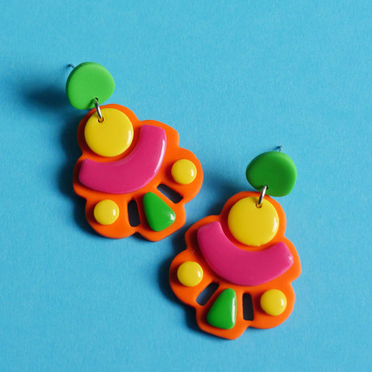 Boucles d'oreilles en forme abstraite, plates de couleur fluo (orange, rose, vert et jaune). L'arrière-plan est un fond bleu clair. 