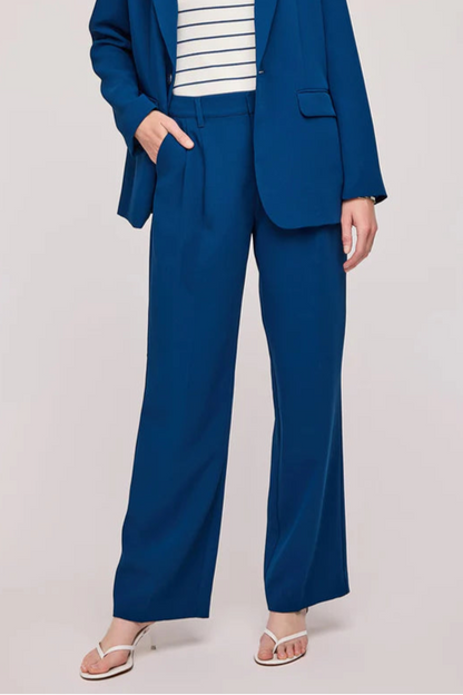 Pantalon bleu marine à la coupe fluide et élégante. Taille haute avec passants pour ceinture, fermeture éclair et bouton discret. Le tissu est légèrement texturé, offrant un tombé impeccable. La coupe droite allonge la silhouette et apporte une touche sophistiquée. Le mannequin porte le pantalon avec un blazer assorti et un haut rayé, ainsi que des sandales à talons.