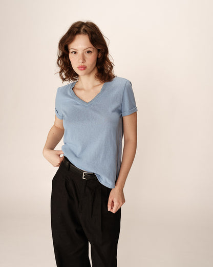 L’image montre une jeune femme portant un T-shirt bleu ciel à col en V, légèrement rentré dans un pantalon noir large.