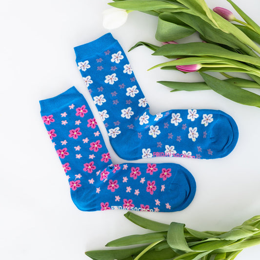 Paire de chaussettes bleu dépareillées. Une chaussette a des petites fleurs roses et l'autre chaussettes des petites fleurs blanches et grises. Les chaussettes sont sur un fond blanc, il y a un bouquet de tulipes posées à côté.