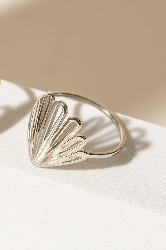 Cette bague à une forme qui rappelle celle d'un coquillage ou des pétales. Sur la photo, on peut voir la version argent et la version dorée. Elles sont déposées sur une pièce en céramique blanche, l'arrière-plan est beige. 