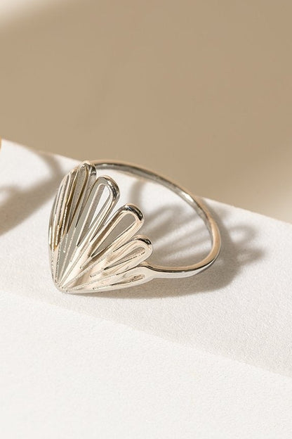 Cette bague à une forme qui rappelle celle d'un coquillage ou des pétales. Sur la photo, on peut voir la version argent et la version dorée. Elles sont déposées sur une pièce en céramique blanche, l'arrière-plan est beige. 