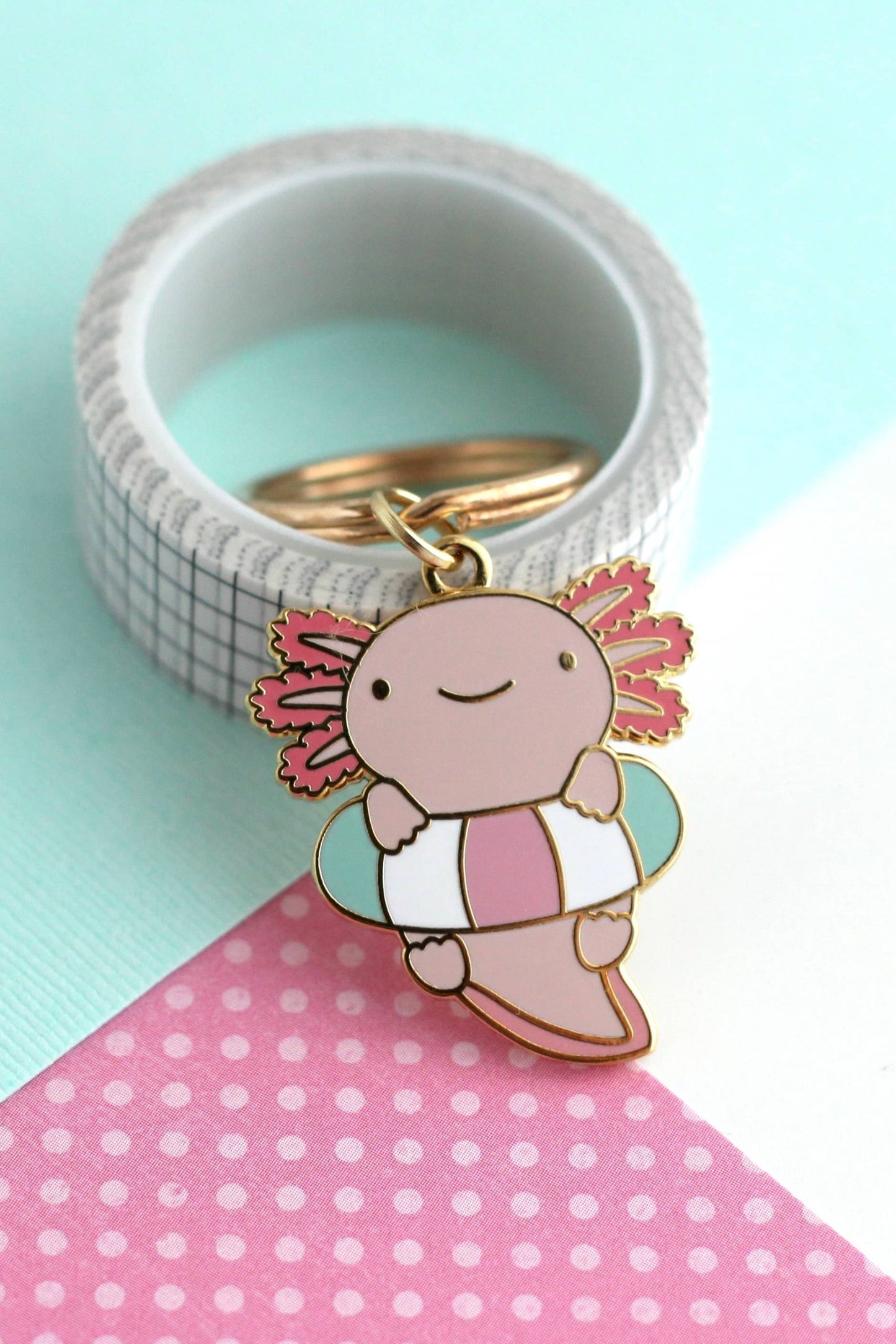 Porte-clés Axolotl Flottant en émail dur doré posé sur fond turquoise et rose à pois, design kawaii pastel.